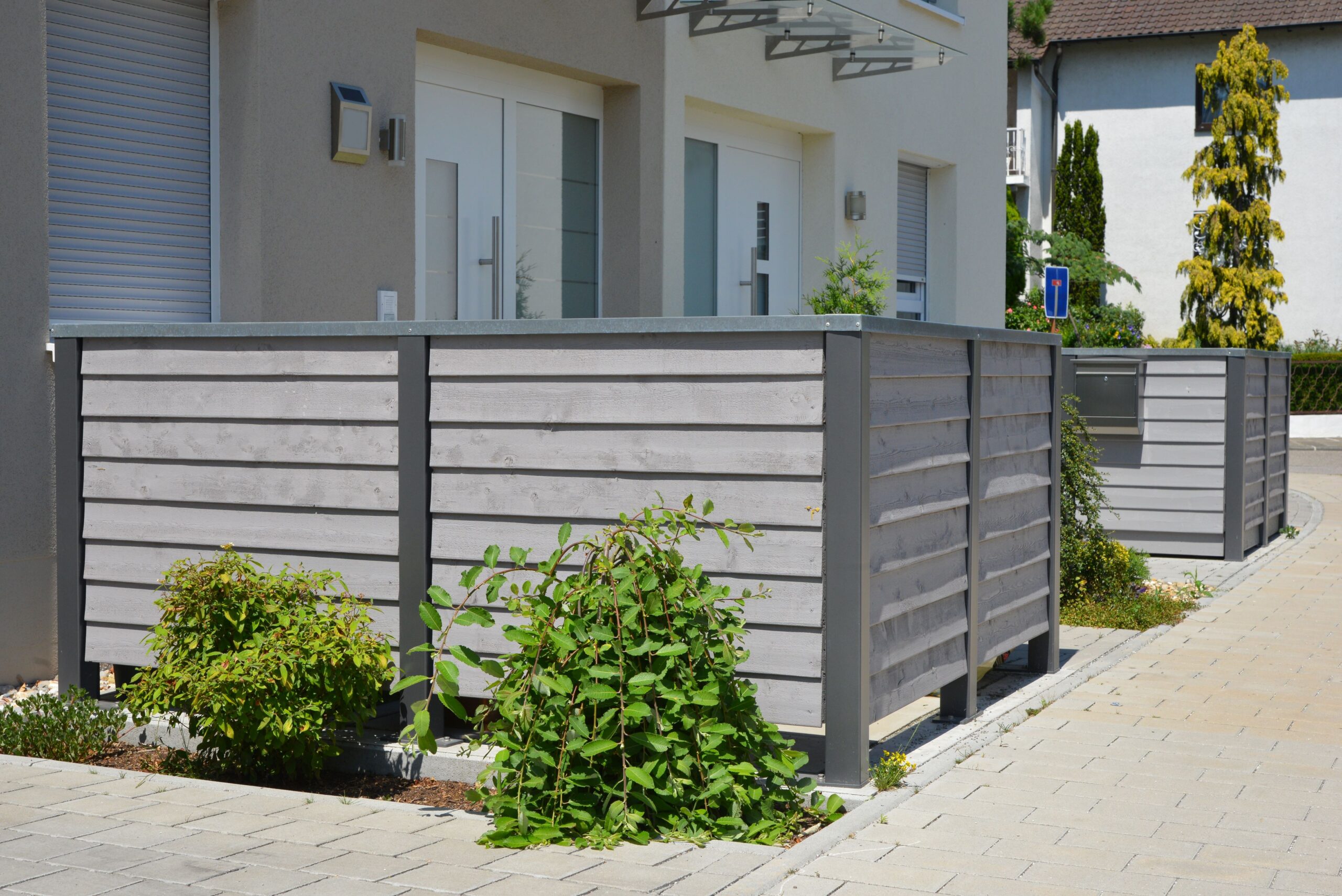 Große, pflegeleichte Mülltonnenverkleidung aus Holzoptik mit Metallrahmen vor modernem Wohnhaus